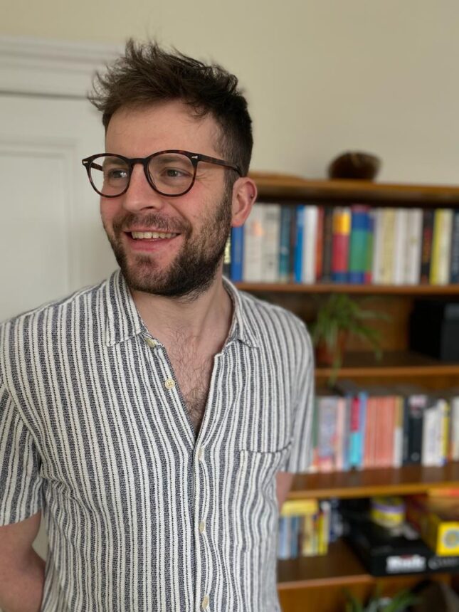 A man wearing glasses and a striped shirt smiling while standing in front of a wooden bookshelf filled with colourful books and a plant.